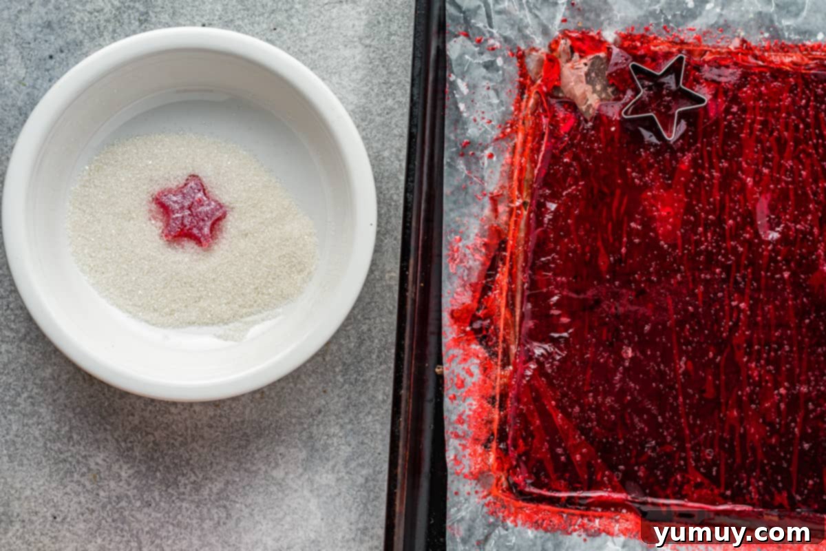 A red star-shaped gumdrop being coated in a bowl of coarse sugar, with a sheet of green gelatin candy in the background.