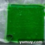 A sheet of green gumdrop candy on parchment paper, with a Christmas tree cookie cutter pressed into one corner, ready for cutting.