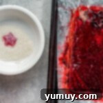A vibrant red star-shaped gumdrop being rolled in a bowl of coarse sugar, with a green gelatin sheet in the background.