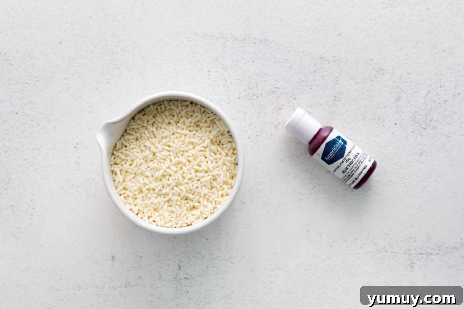 Crafting Custom Colored Sprinkles 7 A small bowl filled with white/plain sprinkles, positioned next to a bottle of vibrant gel food dye, ready for the coloring process.