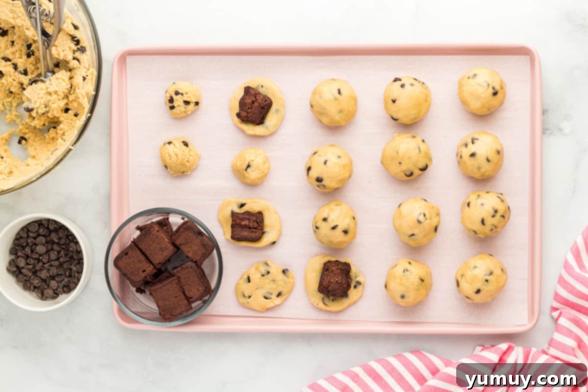 balls of cookie dough arranged on a baking tray, cubes of brownies being placed at the center of some of the cookies