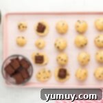 balls of cookie dough arranged on a baking tray, cubes of brownies being placed at the center of some of the cookies