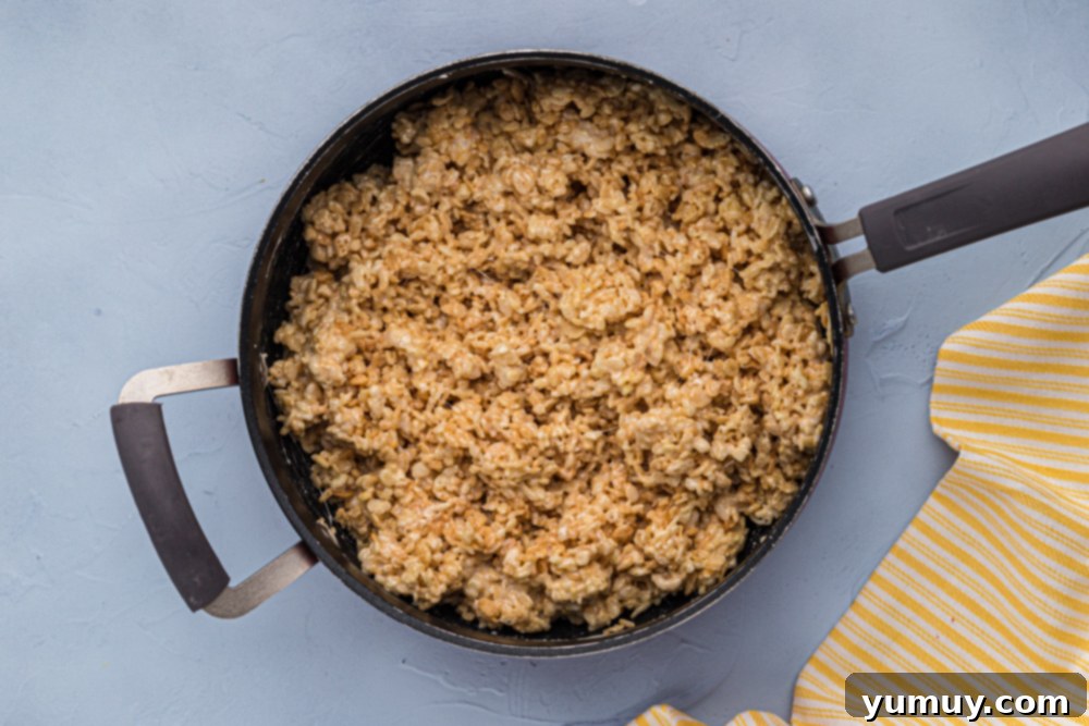 Crispy Marshmallow Squares 7 Brown rice cereal being mixed into a gooey marshmallow mixture in a skillet on a blue background.