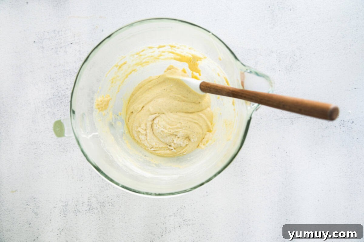 Delicate French Shell Cakes 12 folding madeleine batter in a glass bowl with a rubber spatula.