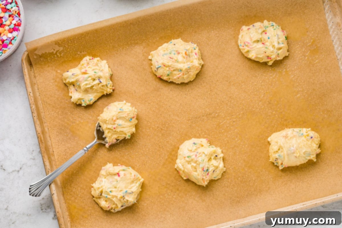 Cake Batter Confetti Cookies 13 Unbaked cookie dough balls, generously dotted with colorful sprinkles, arranged on a parchment-lined baking tray, ready for the oven.