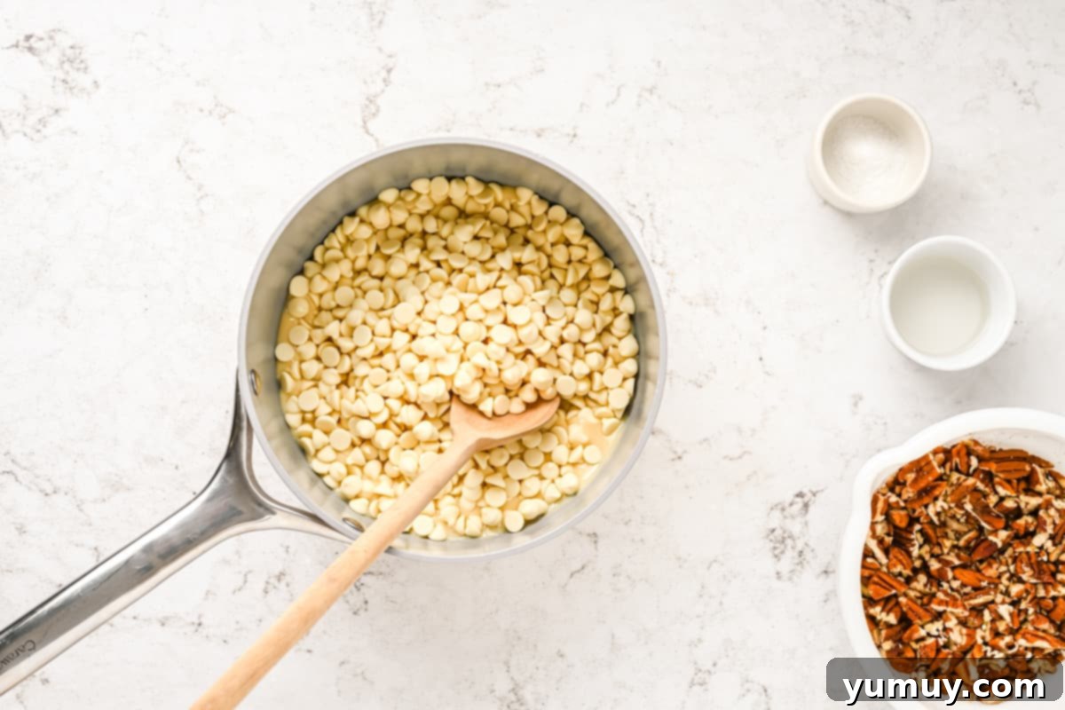 White chocolate chips and sweetened condensed milk melting in a saucepan, being stirred with a wooden spoon.