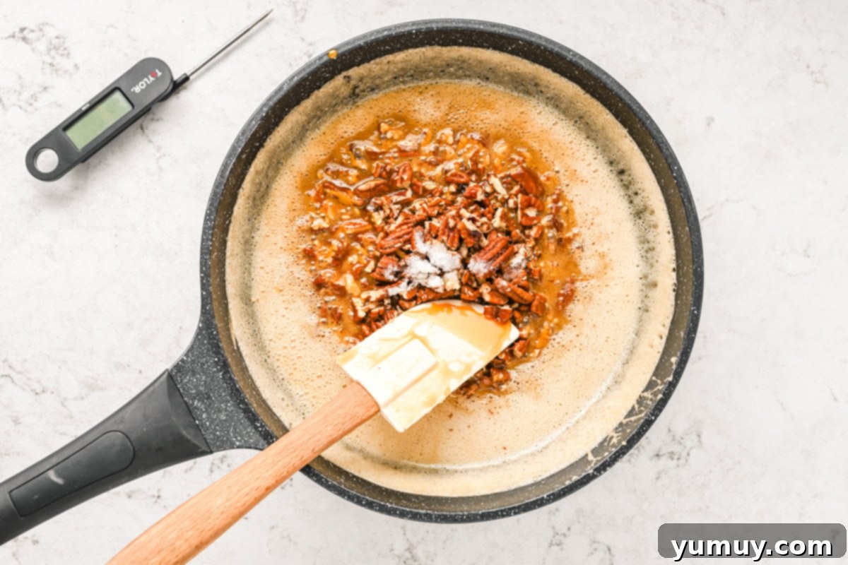 Pecans, vanilla extract, and salt being stirred into the hot praline mixture in a frying pan with a wooden spatula.
