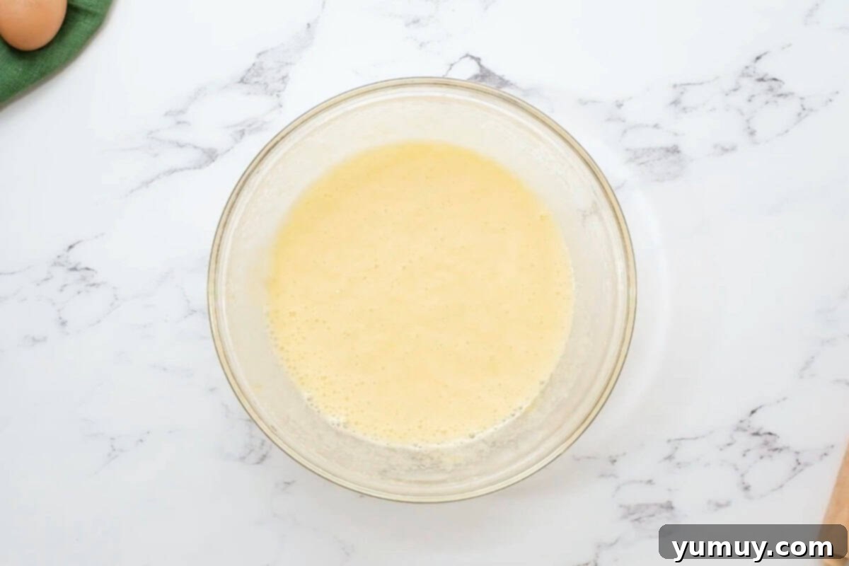 A glass mixing bowl filled with whisked eggs and granulated sugar, forming a light yellow, frothy mixture.