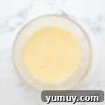 A glass mixing bowl filled with whisked eggs and granulated sugar, forming a light yellow, frothy mixture.