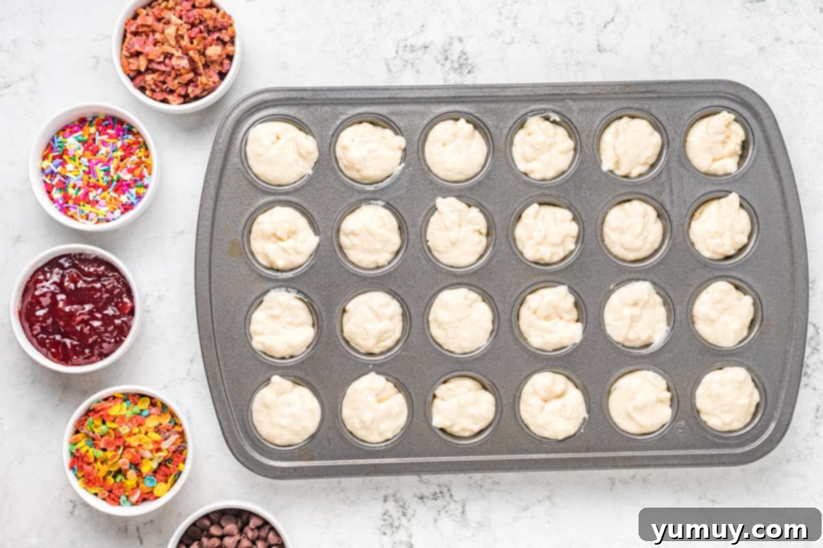 A mini muffin tin filled with pancake batter, with small bowls of various mix-ins like chocolate chips and berries nearby.