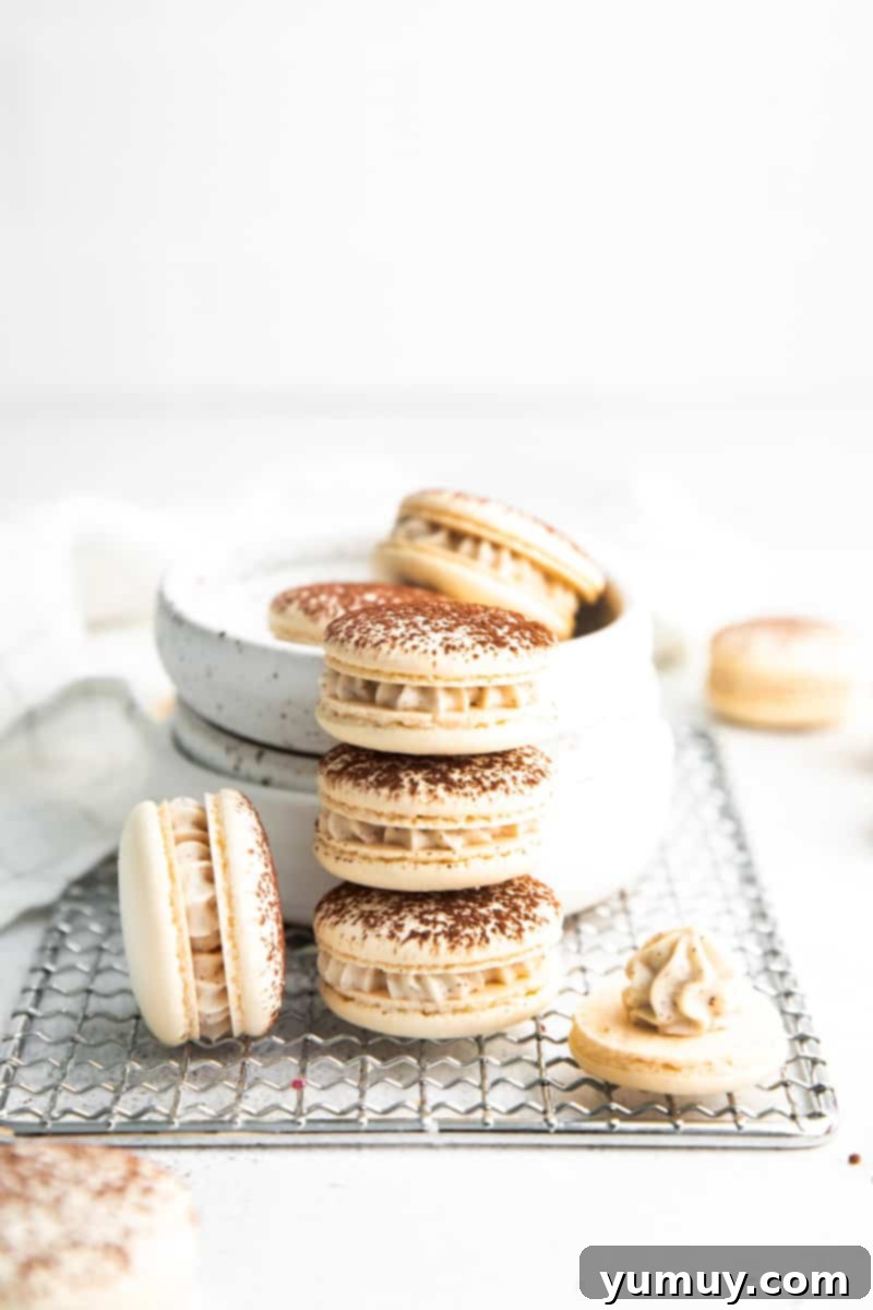 Three perfectly stacked Tiramisu Macarons, with an additional macaron half-filled with frosting, all arranged beside a white bowl overflowing with more macarons. The scene suggests a decadent and delightful dessert experience.
