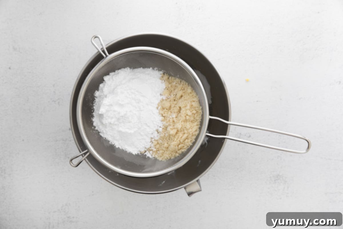 A fine-mesh sieve rests over a stand mixer bowl filled with whipped egg whites, with powdered sugar and almond flour being sifted through, illustrating a key step in macaron preparation.