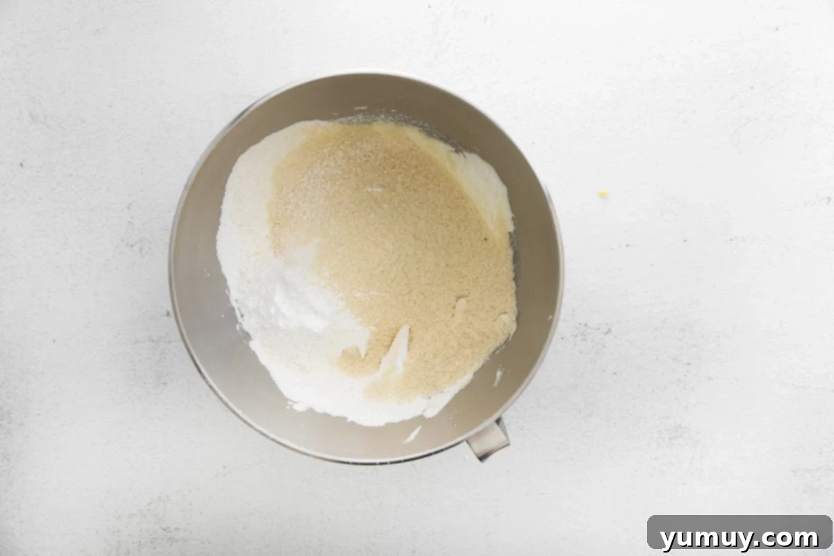 Sifted almond flour and powdered sugar delicately sit atop whipped egg whites in a stainless steel stand mixer bowl, poised for the crucial macaronage folding process.