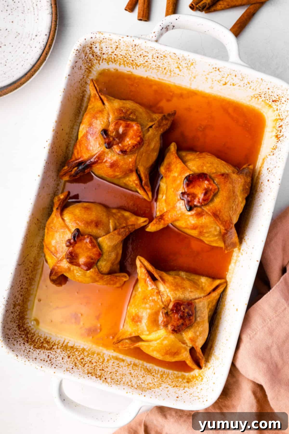Flaky Pie Crust Apple Delights 3 Overhead view of four apple dumplings baking in a pan, drenched in sweet sauce.