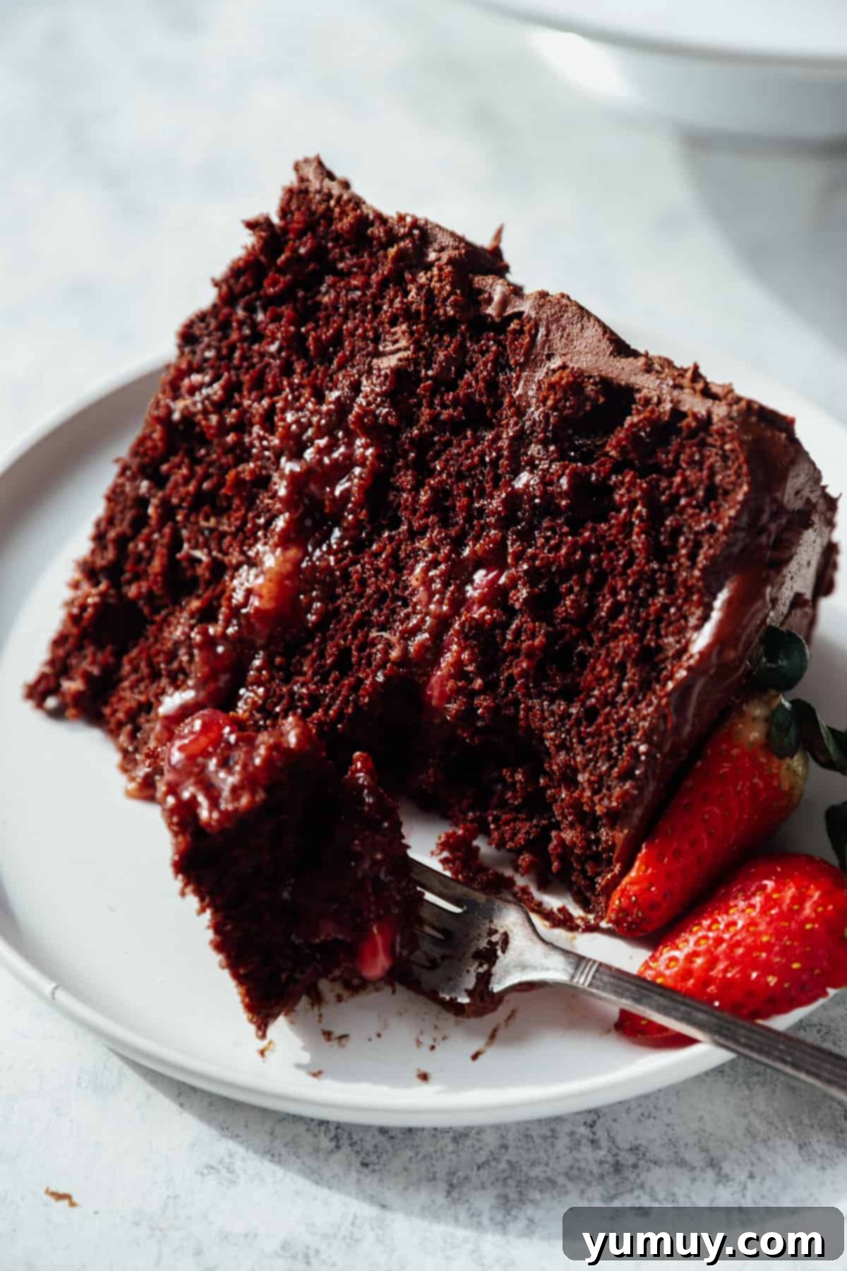 Decadent Chocolate Strawberry Delight 3 A forkful of decadent chocolate strawberry cake on a pristine white plate, with another full slice visible in the background, highlighting the distinct layers of chocolate cake and vibrant strawberry filling.