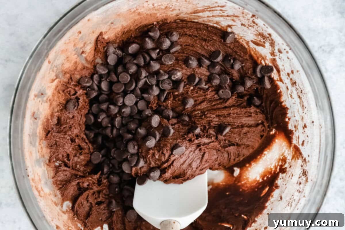 A white rubber spatula folding chocolate chips into cosmic brownie cookie dough in a glass bowl.