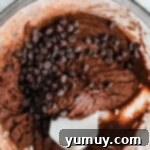 A white rubber spatula folding chocolate chips into cosmic brownie cookie dough in a glass bowl.