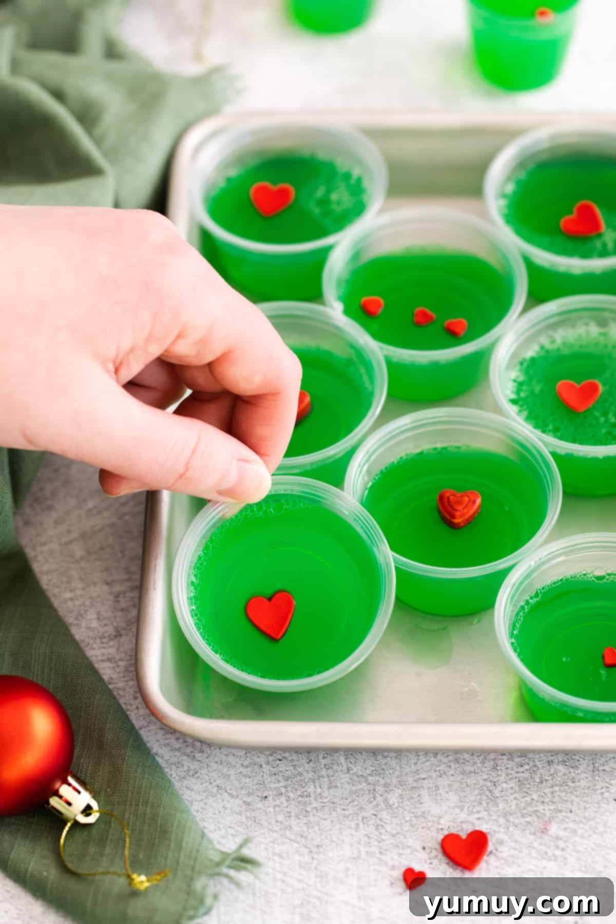A hand reaching for one of eight festive green Grinch jello shots, garnished with red candy hearts, arranged on a rimmed baking sheet.