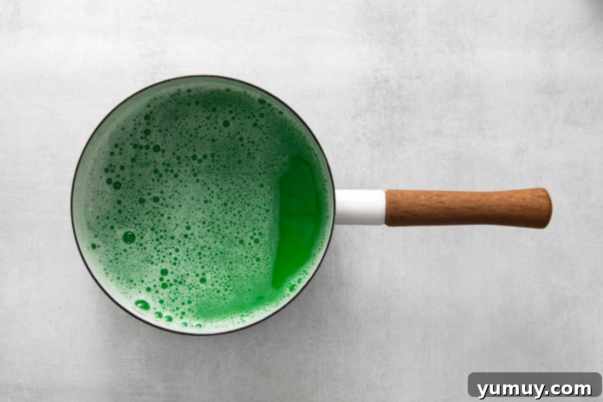 Green food coloring being whisked into the liquid gelatin mixture in a saucepan.