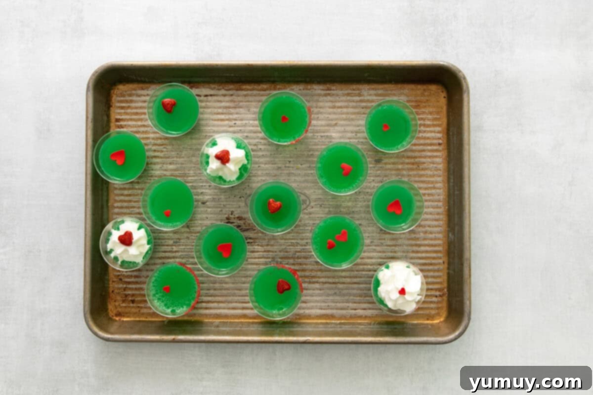 Overhead view of fifteen vibrant green Grinch jello shots, each garnished with a red candy heart, neatly arranged on a rimmed baking sheet.