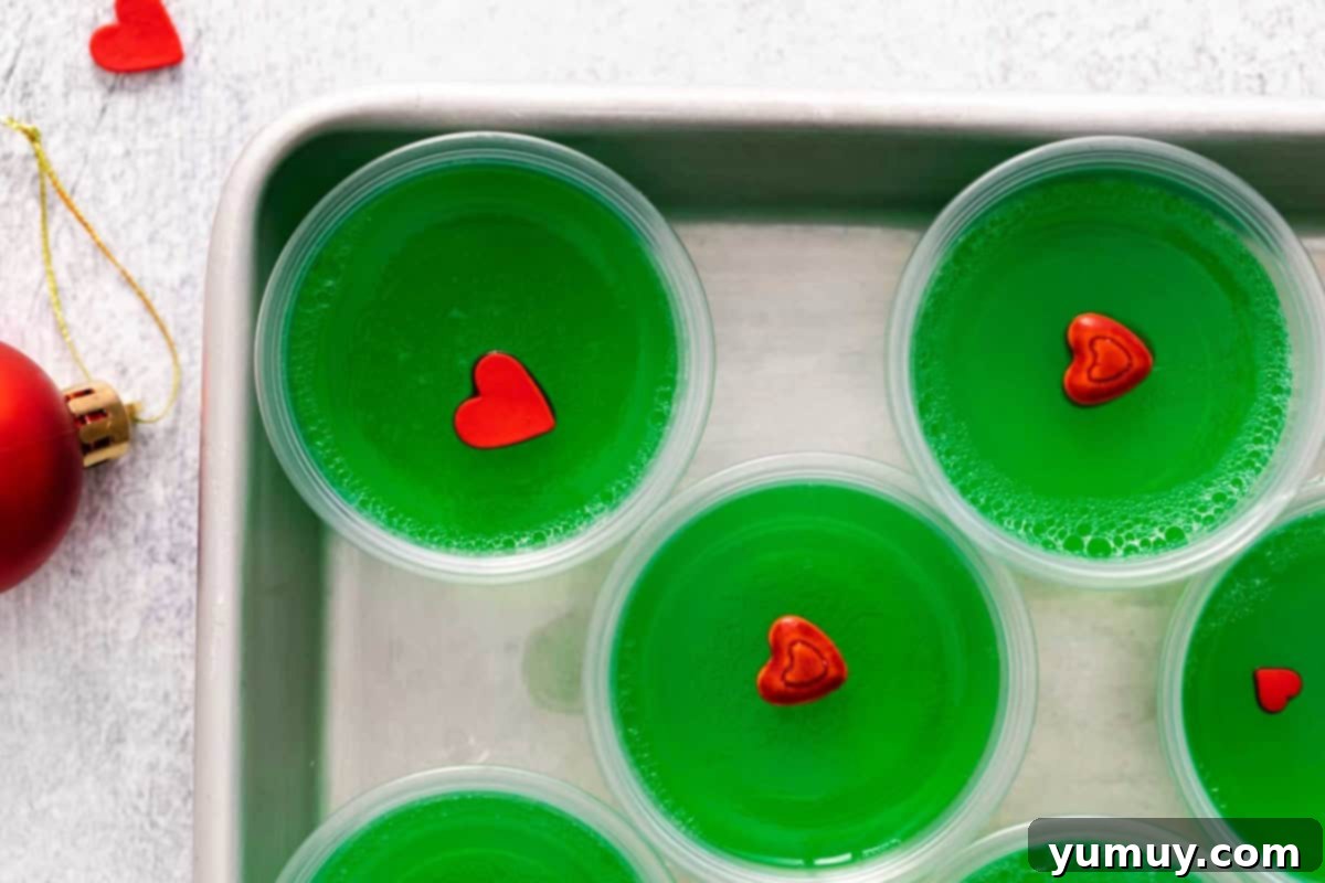 Closeup overhead view of vibrant green Grinch jello shots, each topped with a delicate red candy heart, meticulously arranged on a clean baking sheet.
