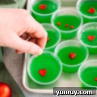 A hand reaching for one of eight festive green Grinch jello shots, garnished with red candy hearts, arranged on a rimmed baking sheet.