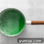 Green food coloring being whisked into the liquid gelatin mixture in a saucepan.