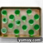 Fifteen unset green Grinch jello shots neatly arranged in individual cups on a rimmed baking sheet.