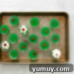 Overhead view of fifteen vibrant green Grinch jello shots, each garnished with a red candy heart, neatly arranged on a rimmed baking sheet.