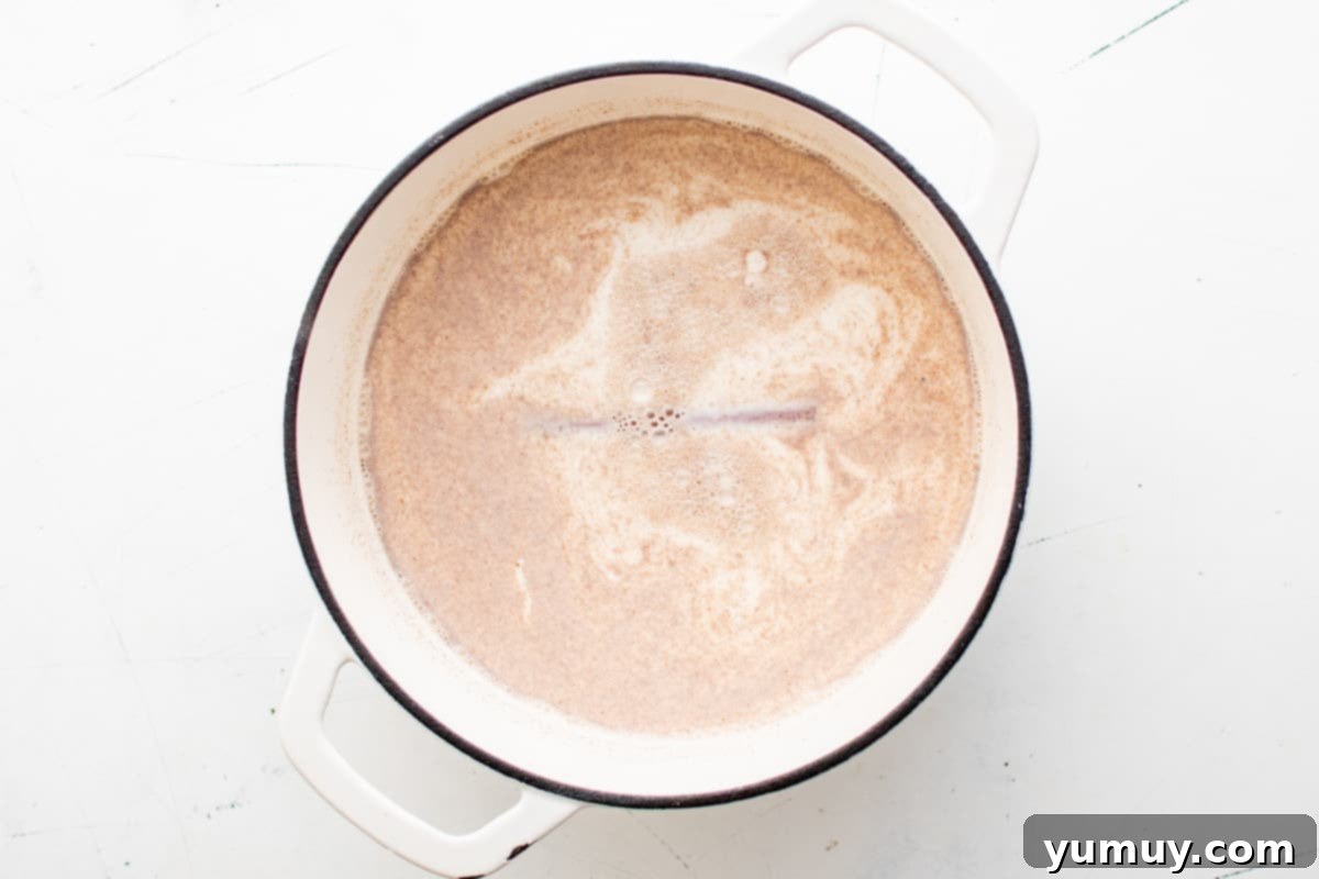 Overhead view of the cinnamon and milk paste being added to a mixture of milk, cream, sugar, and whole cinnamon sticks in a white Dutch oven.