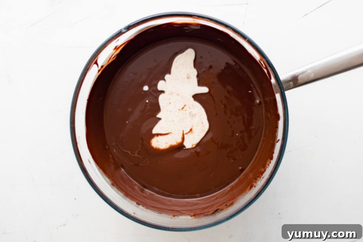 Overhead view of warm cream being poured into melted chocolate in a glass bowl, with a spoon whisking the mixture to temper.