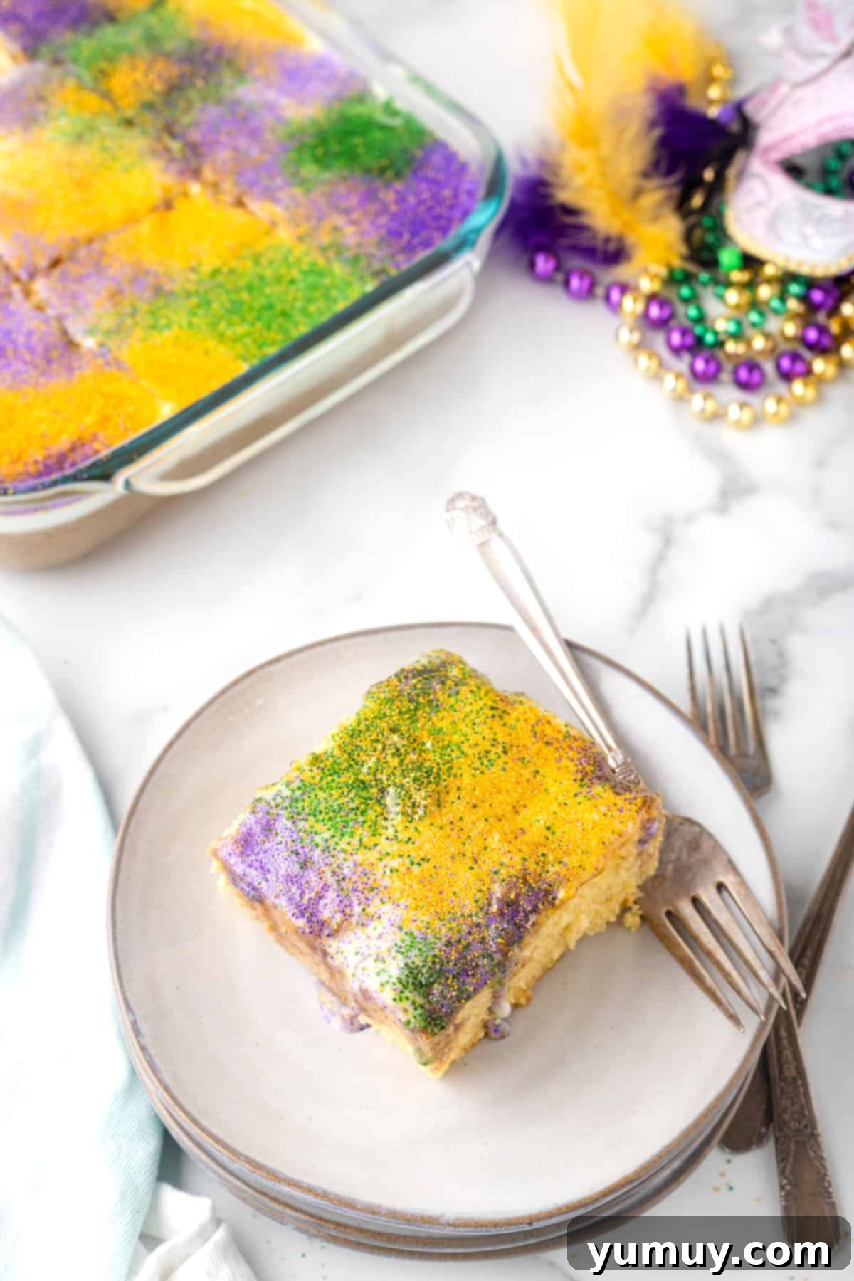 Mardi Gras King Cake Poke 2 A vibrant slice of King Cake Poke Cake on a white plate, showcasing its moist texture and festive purple, green, and gold sprinkles, ready for Mardi Gras celebrations.