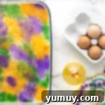 Mardi Gras King Cake Poke 12 The fully decorated King Cake Poke Cake, brightly adorned with generous stripes of green, gold, and purple decorating sugars over its white glaze.