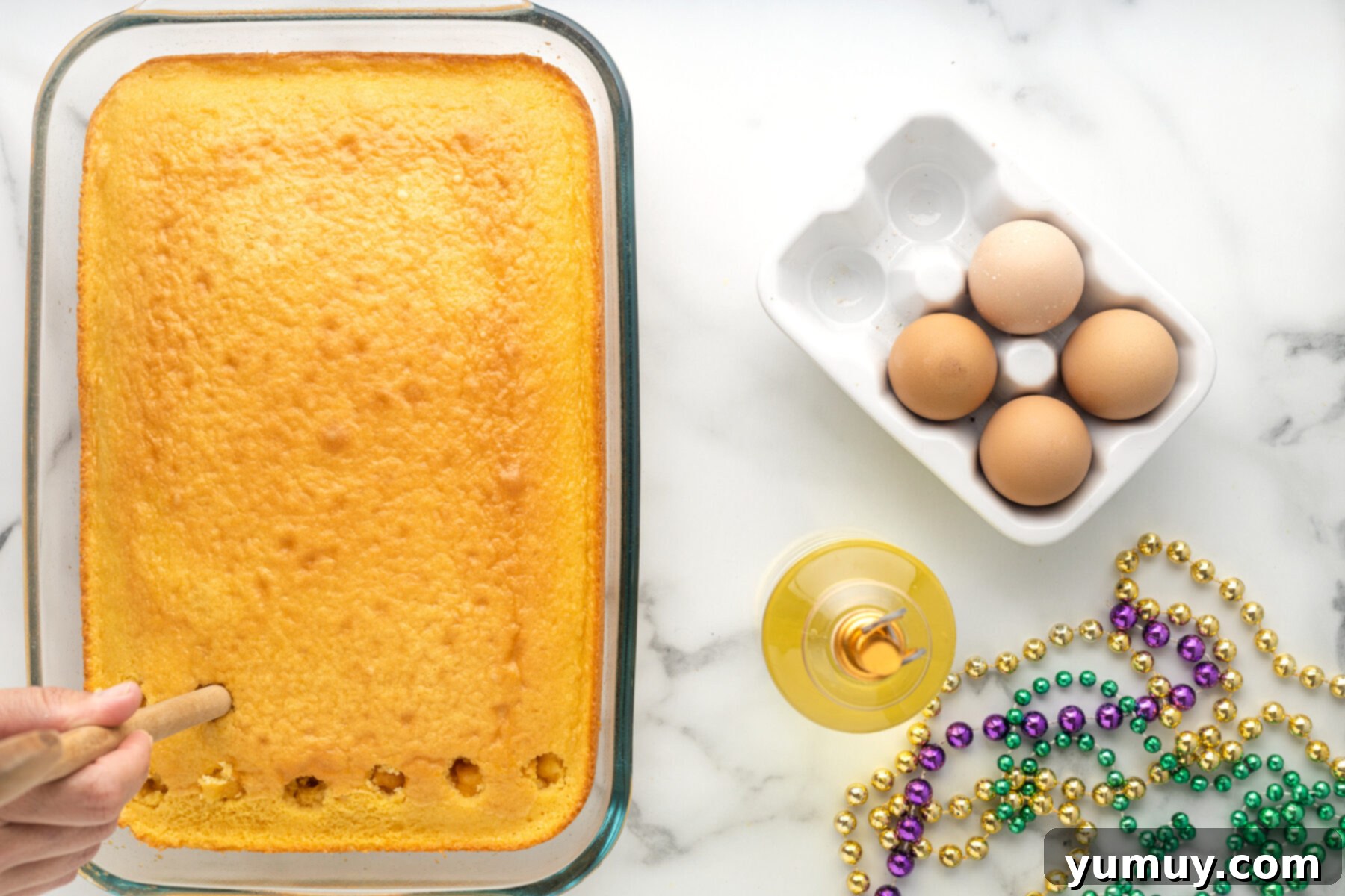 Mardi Gras King Cake Poke 14 A hand using the handle of a wooden spoon to carefully poke holes into the surface of a baked yellow cake, creating depressions for the pudding.