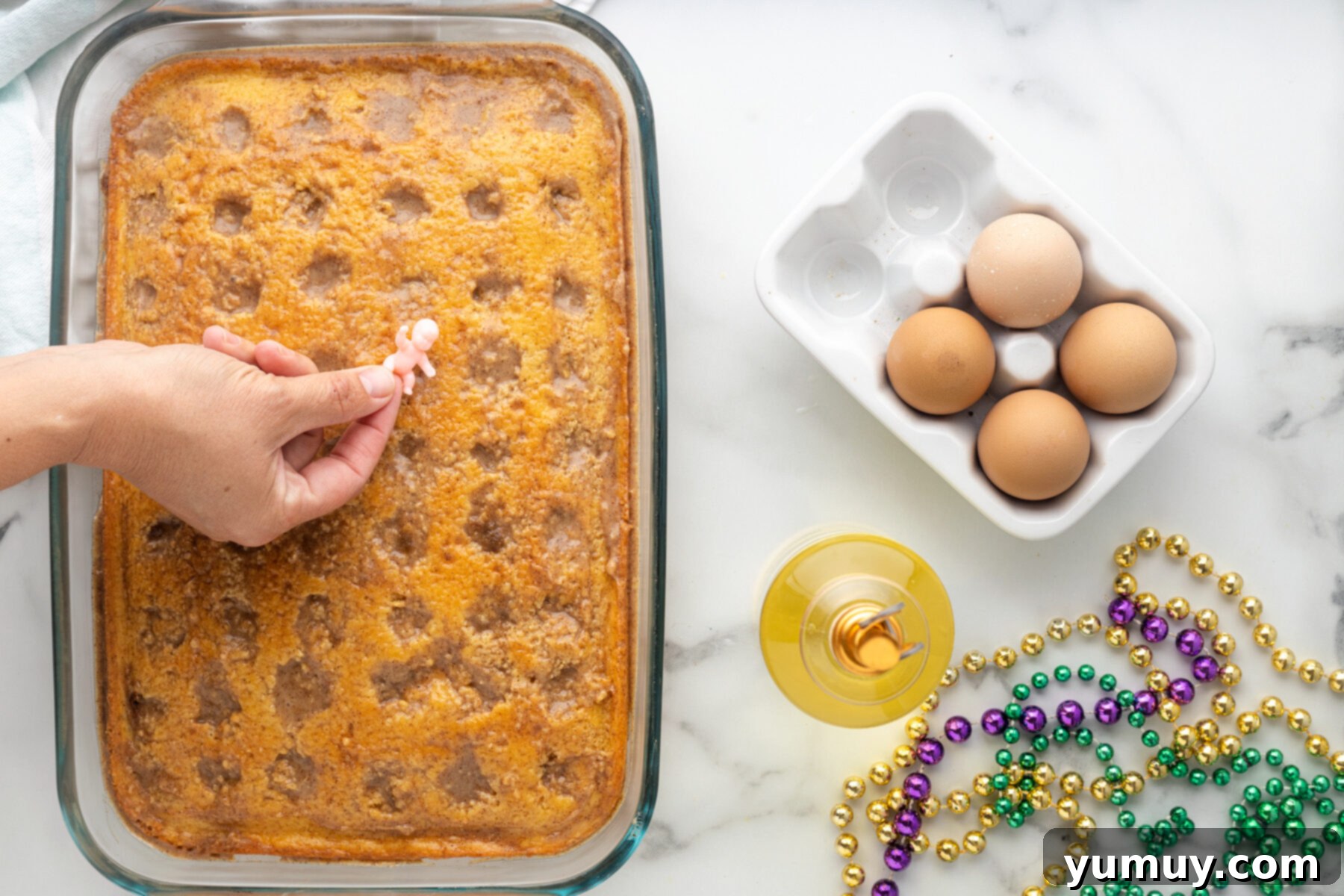 Mardi Gras King Cake Poke 16 A hand gently placing a tiny plastic baby figurine into a set King Cake Poke Cake, ensuring it is hidden within the dessert for the traditional Mardi Gras game and surprise.