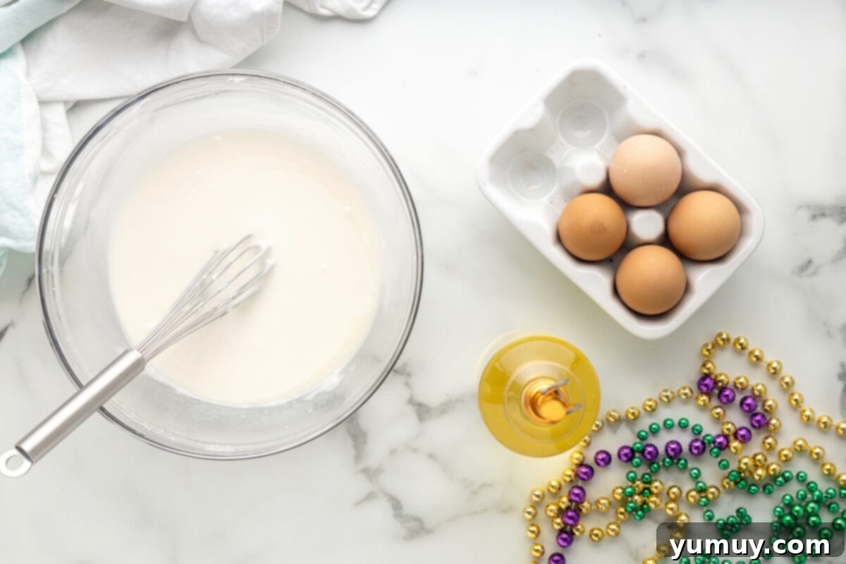Mardi Gras King Cake Poke 17 A perfectly smooth, white glaze for the King Cake Poke Cake, resting in a clear glass bowl with a whisk, showing its ideal pourable consistency.