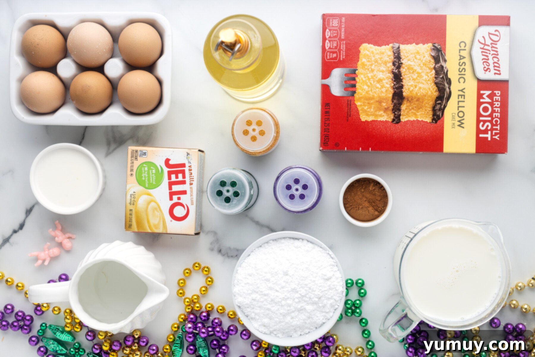 Mardi Gras King Cake Poke 3 An overhead view of the organized ingredients needed for King Cake Poke Cake, including a yellow cake mix box, vanilla pudding, milk, eggs, vegetable oil, cinnamon, powdered sugar, and colorful decorating sugars, laid out on a clean surface.
