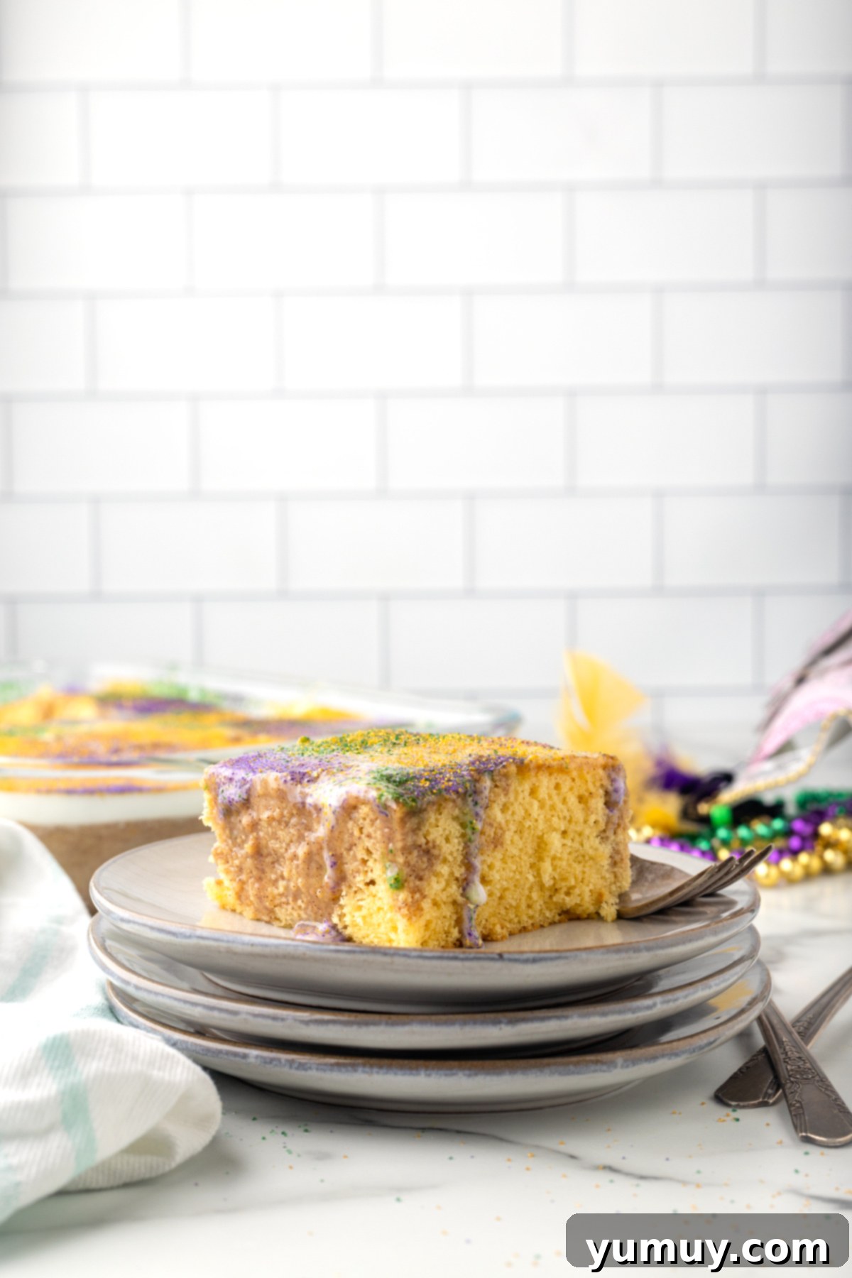 Mardi Gras King Cake Poke 4 A delicious side view of a slice of King Cake Poke Cake, featuring distinct layers of yellow cake, cinnamon pudding, and white glaze topped with colorful sprinkles, presented on a stack of white plates.