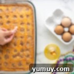 Mardi Gras King Cake Poke 9 A hand carefully placing a tiny plastic baby figurine into a set King Cake Poke Cake, ensuring it is hidden within the dessert for the traditional Mardi Gras game.
