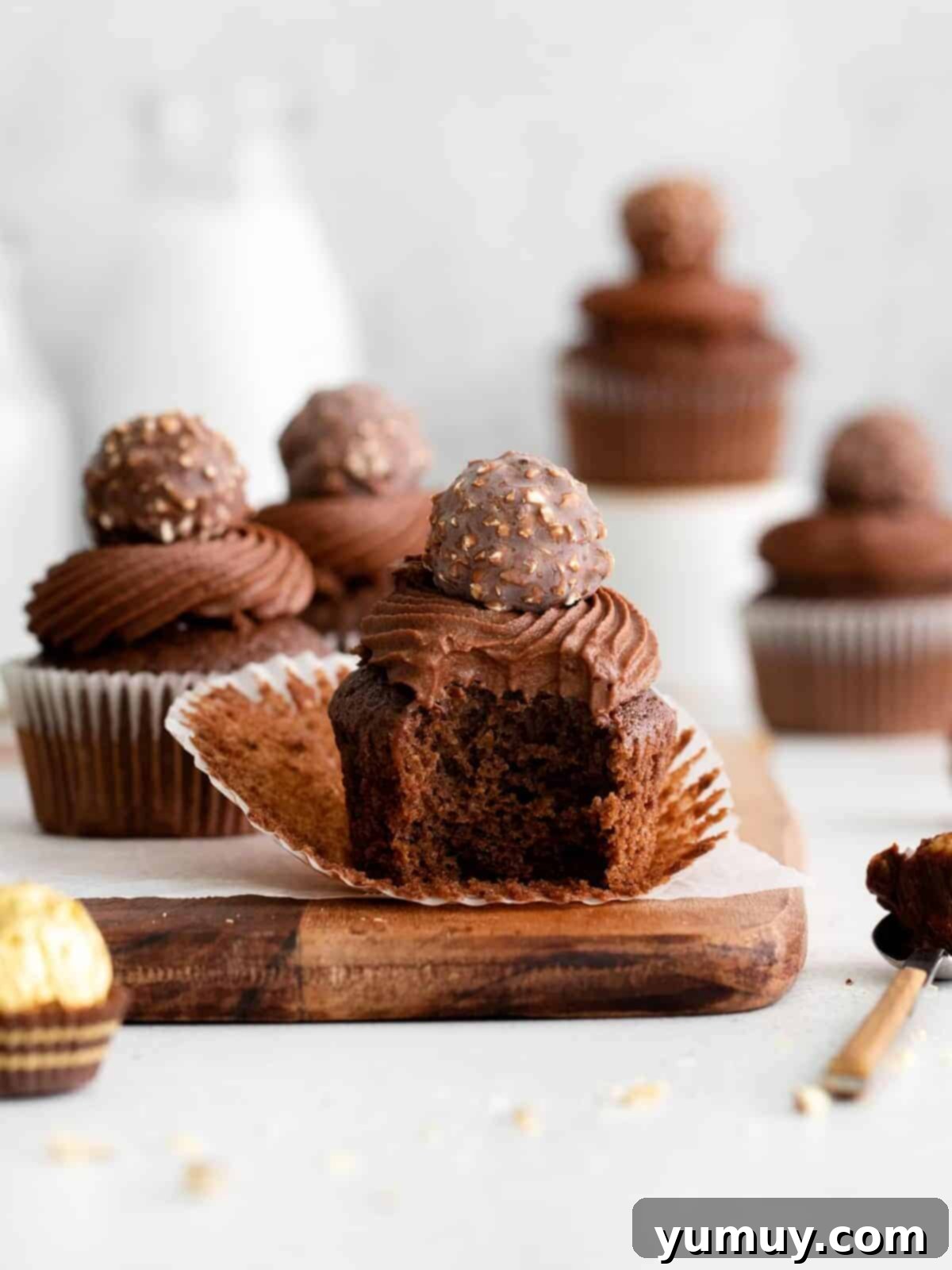 An unwrapped and bitten Ferrero Rocher cupcake on a rustic wooden cutting board, showcasing its rich chocolate cake, creamy hazelnut filling, and fluffy ganache frosting, with a whole Ferrero Rocher candy on top.