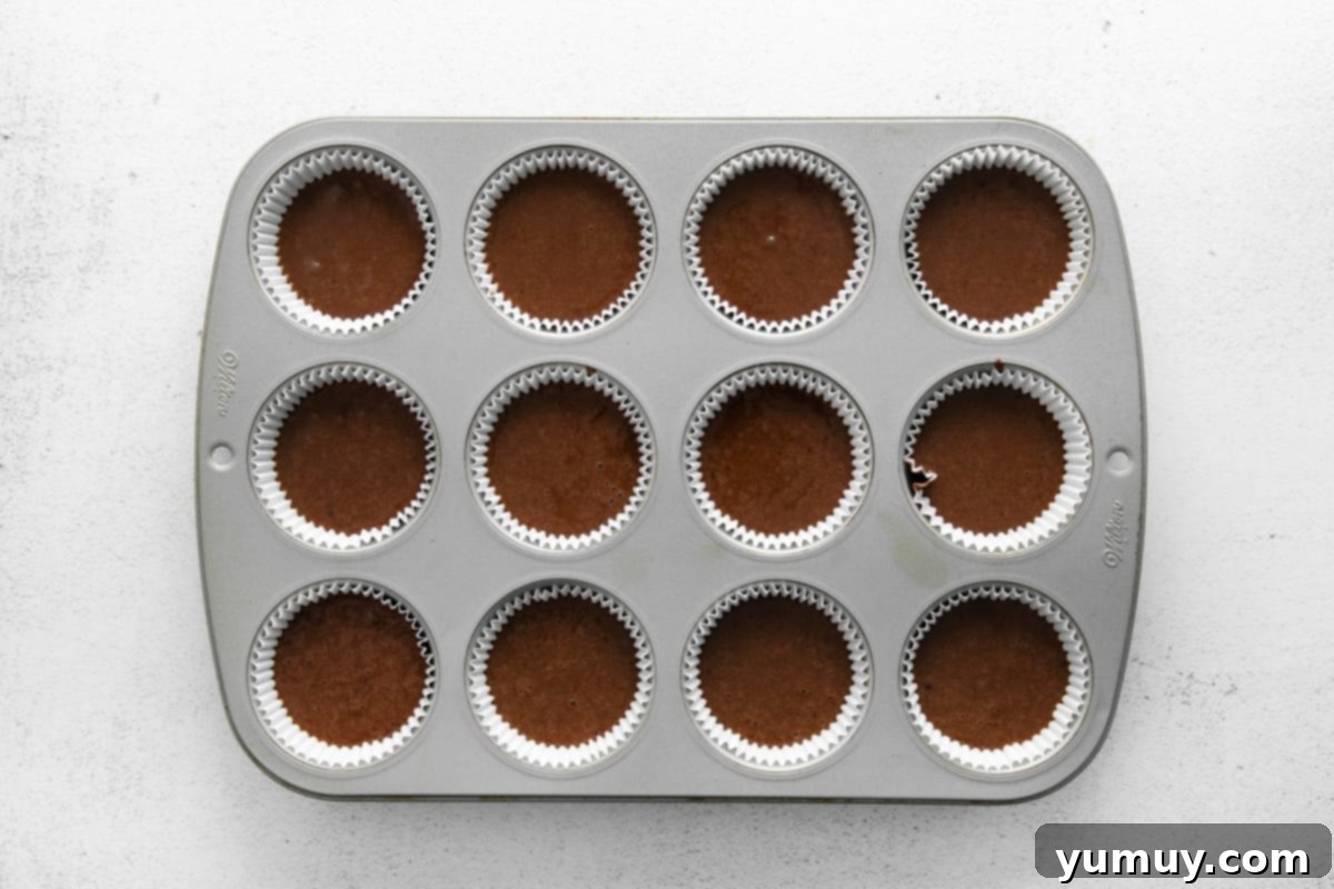 A 12-count cupcake tin, with each liner filled evenly with chocolate cupcake batter, poised for baking.