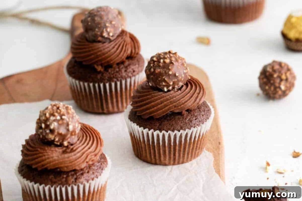 Three perfectly crafted Ferrero Rocher cupcakes displayed on a wooden cutting board. Each cupcake features rich chocolate cake, fluffy chocolate-hazelnut ganache, and is adorned with a Ferrero Rocher candy, highlighting their elegant presentation.