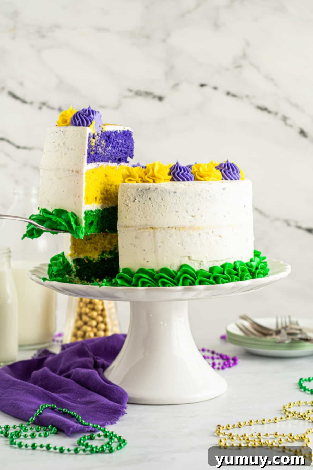 Mardi Gras Birthday Fiesta 4 A side view of a perfect slice of Mardi Gras birthday cake being lifted from the larger cake on a white cake stand, highlighting the colorful layers and frosting.