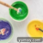 Mardi Gras Birthday Fiesta 7 Purple, green, and yellow Mardi Gras birthday cake batter in separate white bowls with rubber spatulas, showcasing their vibrant distinct colors.