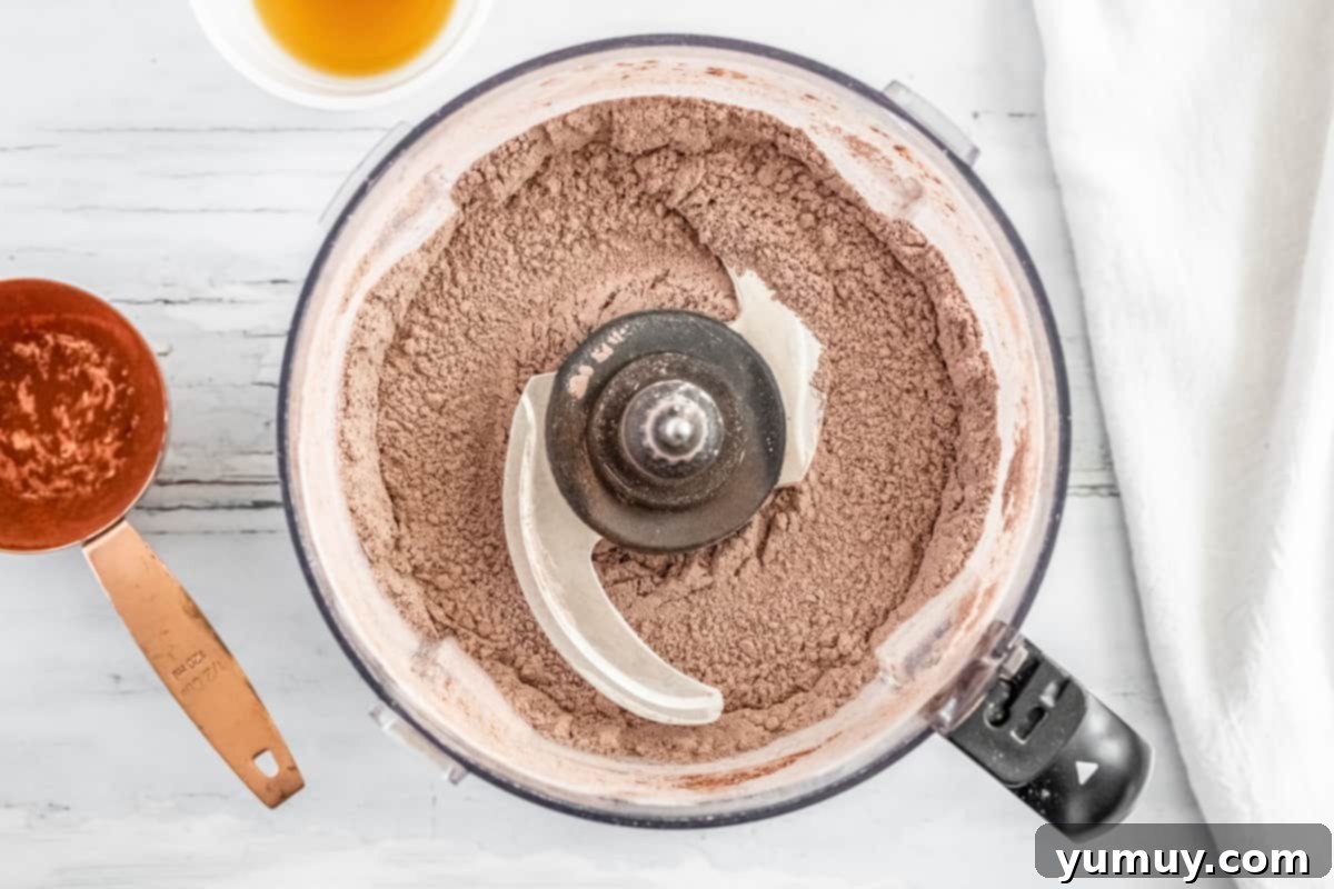 Close-up of mixed dry ingredients for Thin Mints in a food processor, displaying the cocoa powder, flour, and sugar blend before liquid is added.