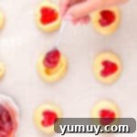 Heart Kiss Cookies 12 a hand using a spoon to add jam to heart thumbprint cookies.