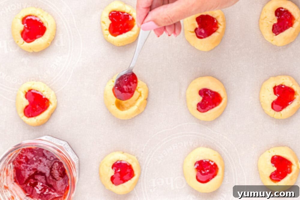 Heart Kiss Cookies 21 A hand carefully spooning vibrant strawberry jam into the heart-shaped indentations of cookie dough balls on a baking sheet.