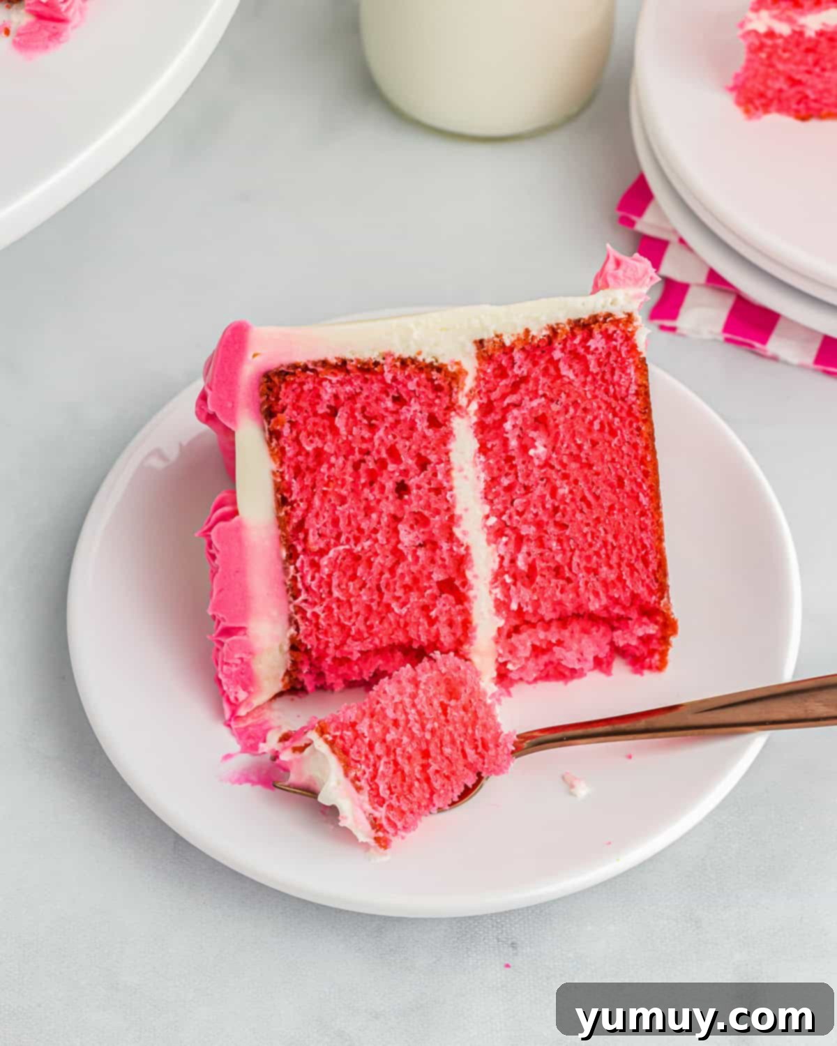 A partially eaten slice of neon pink velvet cake on a white plate, served with a fork, showcasing its vibrant color and smooth cream cheese frosting.