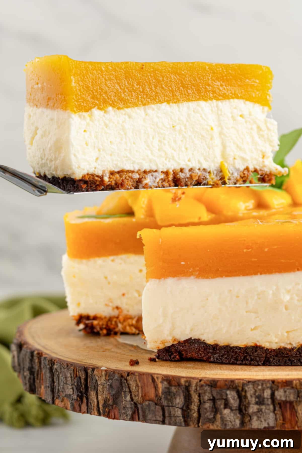 A perfectly sliced piece of no-bake mango cheesecake resting on a rustic wooden plate, garnished with fresh mint leaves and a vibrant mango cube, highlighting its creamy texture and rich mango jam topping.