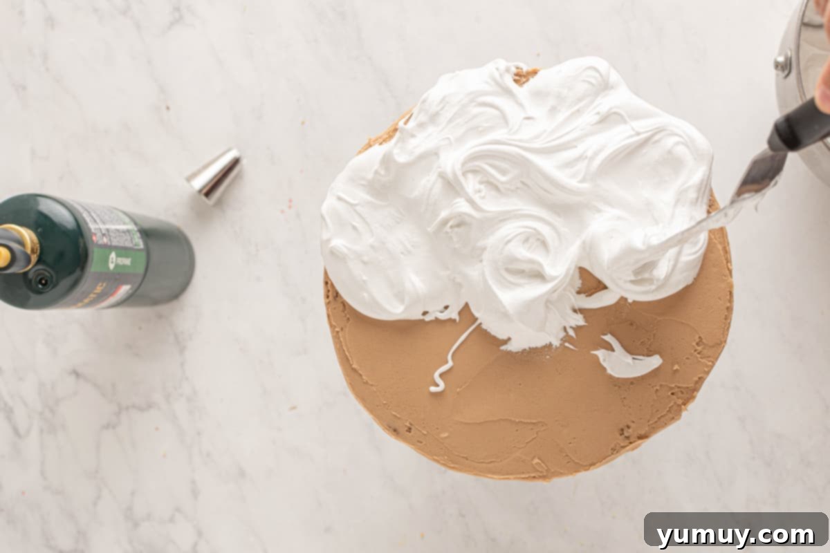 meringue being swirled over a frosted smores cake on a cake stand.
